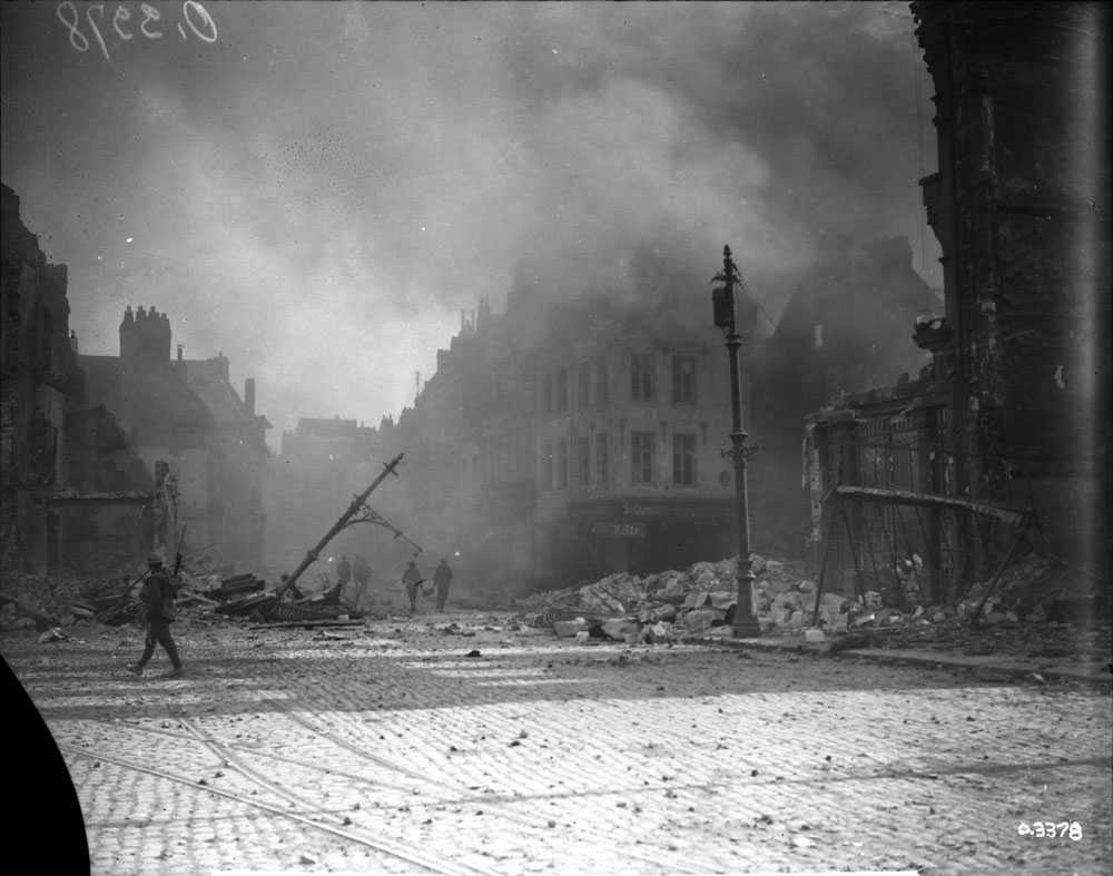 Black and white photograph. Across an abandoned city square, filled with rubble, several individual soldiers can be seen walking. Smoke fills the air.